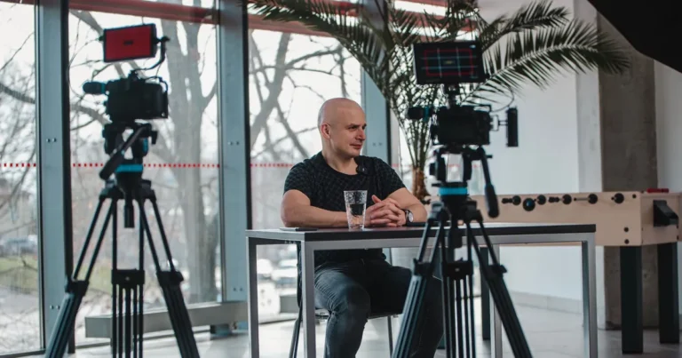 Eugenijus Mačiukas interviu filmavimo metu prie stalo, priekyje dvi kameros ant trikojų