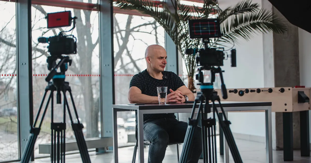 Eugenijus Mačiukas interviu filmavimo metu prie stalo, priekyje dvi kameros ant trikojų