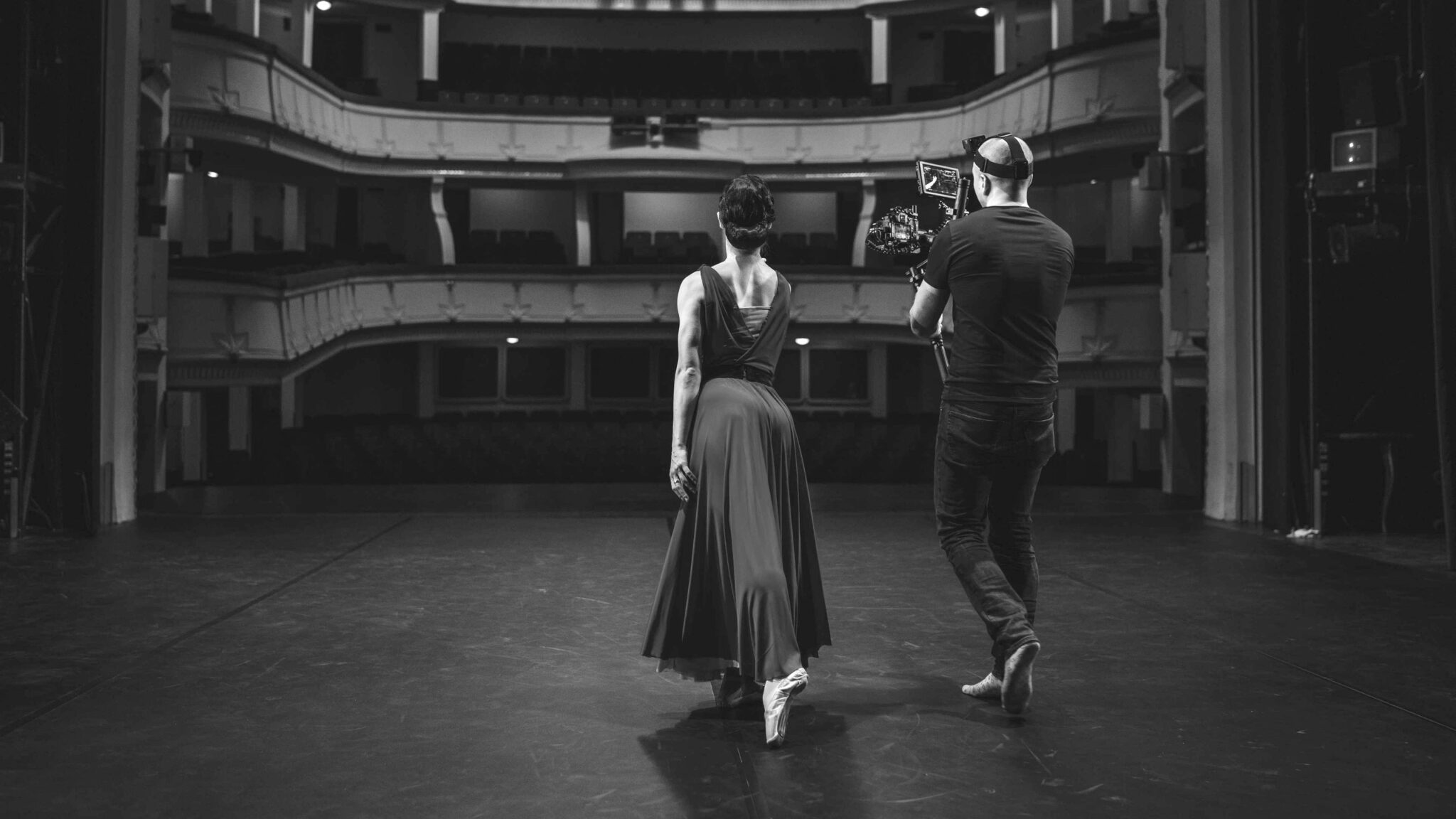 Renginio filmavimas teatro scenoje: operatorius su kamera ir artistė, fone tuščia salė su balkonais